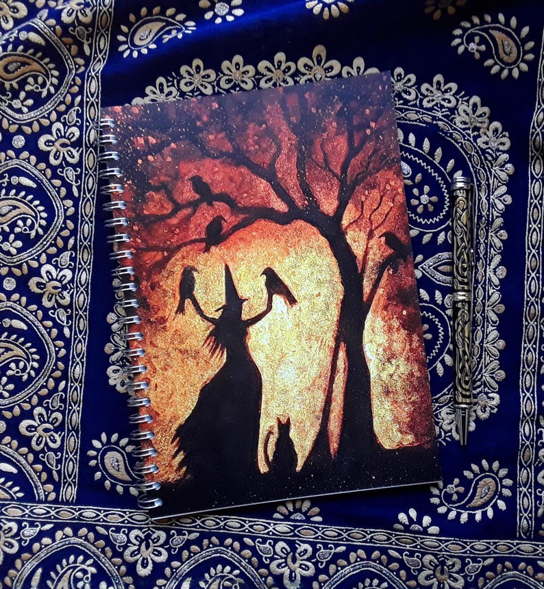 May include: Spiral-bound notebook featuring a witch silhouette with crows and a cat under a tree against a sunset backdrop. A decorative pen rests beside the notebook on a blue patterned fabric.
