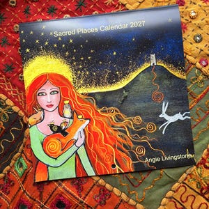 Puede incluir: Un calendario de pared cuadrado titulado Sacred Places Calendar 2027 de Angie Livingstone. La portada presenta una pintura de una persona con largo cabello rojo sosteniendo un zorro, un b&uacute;ho y un p&aacute;jaro, bajo un cielo estrellado con una colina y una liebre.