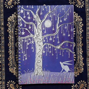 May include: Spiral-bound notebook with a whimsical design. The cover features a silver tree adorned with hanging stars against a deep blue background. A white rabbit sits below the tree. The notebook is on a dark blue fabric.