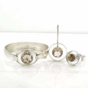 Peut inclure: Un ensemble de trois bijoux en argent avec des pierres de quartz fum&eacute;. L'ensemble comprend un bracelet, un pendentif et une bague. Chaque pi&egrave;ce pr&eacute;sente un design circulaire avec une seule pierre au centre.