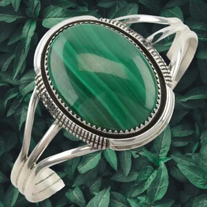 May include: A silver cuff bracelet with a large, oval malachite stone. The green stone is set in a detailed silver bezel with a black border. The bracelet has a split design with decorative silver accents.