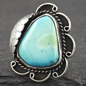 May include: A silver-toned ring featuring a large, light blue turquoise stone. The stone is set in a decorative frame with a leaf design on one side and a scalloped edge. The ring has a vintage or Southwestern style.