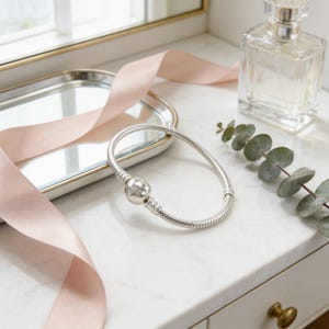 a silver bracelet with a round charm, a clear perfume bottle, and a pink ribbon on a white marble countertop.