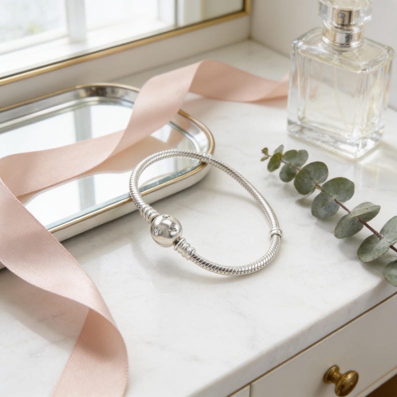 a silver bracelet with a round charm, a clear perfume bottle, and a pink ribbon on a white marble countertop.