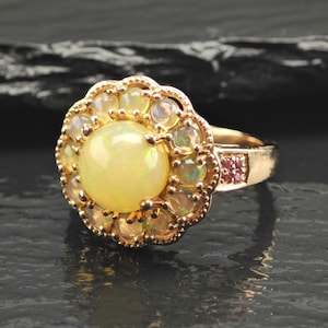 May include: A gold ring featuring a large, round, cabochon-cut opal center stone surrounded by a halo of smaller, round opal stones. The band includes a small cluster of tiny, deep pink gemstones set into the side of the metal setting.