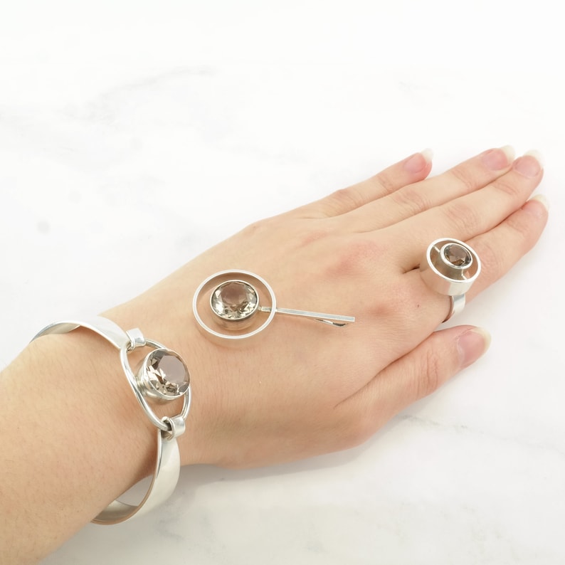 Peut inclure: Un ensemble de trois bijoux en argent avec une pierre pr&eacute;cieuse en quartz fum&eacute;. L'ensemble comprend un bracelet, une bague et une broche. Chaque pi&egrave;ce a un design circulaire avec une pierre pr&eacute;cieuse au centre.
