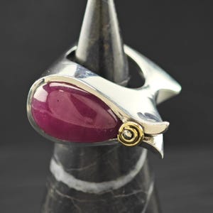 May include: A silver ring featuring a large, oval, deep red gemstone. The ring has a unique, modern design with a gold accent. The ring is displayed on a black and grey stand.