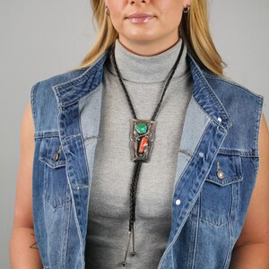 May include: A bolo tie featuring a rectangular silver-toned metal centerpiece with etched detailing, a round green stone, and an elongated orange stone. The centerpiece is attached to a black braided leather cord with silver-toned metal tips.