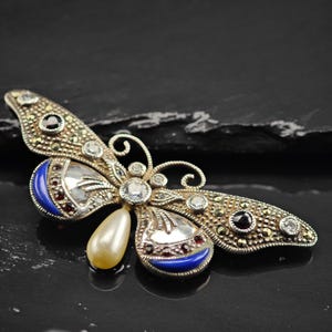 May include: A silver-toned butterfly brooch with marcasite and gemstone accents. The brooch features a pearlescent drop, blue enamel details, and clear and black gemstones. The wings have intricate detailing and a curved antenna design.
