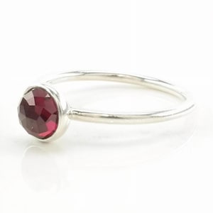 May include: A silver ring featuring a single, round, faceted deep red gemstone set in a smooth metal bezel. The band is thin, polished, and circular, showcasing a minimalist design with a reflective surface and a vibrant, dark crimson stone.