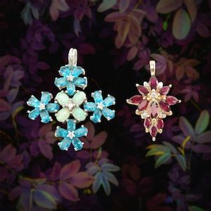 May include: Two floral pendant necklaces. One pendant is silver with blue and white gemstones. The other pendant is gold with red gemstones and small diamonds. The background is a dark purple floral pattern.