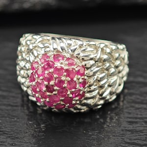 May include: A silver ring featuring a cluster of small, round, bright pink gemstones at the center. The ring's band has a textured, woven design. The gemstones are tightly packed, creating a focal point.