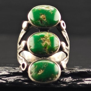 May include: A silver ring featuring three vertically aligned oval green stones with natural brown matrix inclusions. The metal band has a polished finish with small circular accents on the sides, framing the stones in a stacked vertical arrangement.