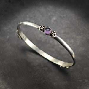 Può includere: Un bracciale rigido in argento con una fascia liscia e lucida e un'incastonatura circolare centrale. L'incastonatura contiene una piccola pietra preziosa di ametista rotonda e viola, affiancata da due decorazioni metalliche a spirale.
