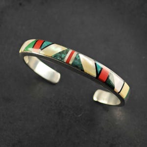 May include: A silver cuff bracelet featuring a geometric inlay pattern of white mother-of-pearl, red coral, and green turquoise segments separated by thin black lines. The metal band has a polished finish and an open back design.