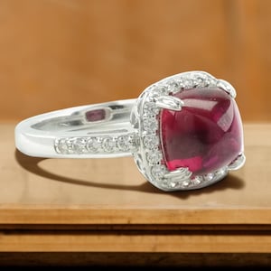 Puede incluir: Un anillo de plata con una gran gema roja oscura de corte cuadrado, rodeada por un halo de gemas claras más pequeñas. La banda del anillo también está adornada con pequeñas gemas claras, creando un efecto brillante. Sobre un fondo de madera cálida.