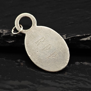 May include: A silver-toned oval pendant featuring the engraved letters R.P.P. in a serif font. The pendant has a smooth, slightly weathered surface and includes a small circular jump ring attached to the top for hanging or connecting to a chain.