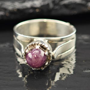 May include: A silver ring with a large, round, deep pink gemstone at the center. The gemstone is set in a decorative bezel. The ring band has a textured design. The ring is likely made of sterling silver.