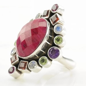 May include: A silver ring featuring a large, oval, faceted deep pink gemstone at the center. The ring is surrounded by smaller, round and square-cut accent stones in various colors, including red, purple, green, and light blue, set in silver.