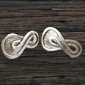 Puede incluir: Dos pendientes plateados con forma de símbolo de infinito abstracto. Cada pendiente presenta una superficie texturizada y martillada en el cuerpo principal y un diseño liso y curvado. Los pendientes se muestran sobre una superficie de madera oscura.
