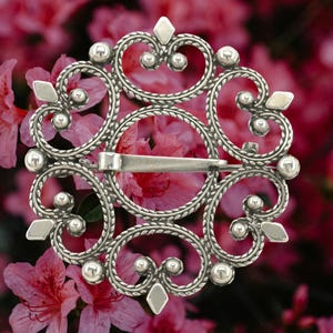May include: A silver-toned brooch with an intricate, openwork design. The brooch features a circular shape with swirling patterns, beaded accents, and diamond-shaped and round embellishments. A clasp is visible on the back. The background is a blurred image of pink flowers.