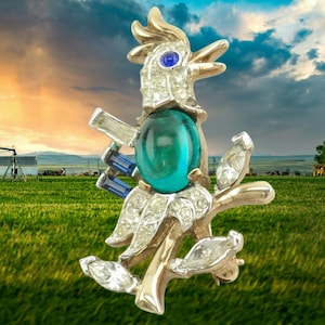 Puede incluir: Broche vintage con forma de gallo, con un gran cabujón de color verde azulado y pedrería transparente y azul. El gallo dorado está posado sobre una rama con detalles de pedrería transparente. El broche está sobre un campo verde y un cielo nublado.