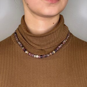 Op de afbeelding: Een veelkleurige kralenketting, met roze, paarse en witte kralen, wordt getoond tegen een bruine coltrui. De ketting bestaat uit kleine, ronde kralen en zit dicht op de hals.