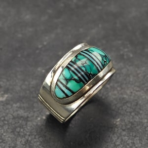 Puede incluir: Un anillo de plata con una incrustación central de piedra turquesa con vetas negras y finas rayas horizontales negras. La banda de metal tiene un acabado pulido con sutiles detalles ranurados a lo largo de los lados del engaste.