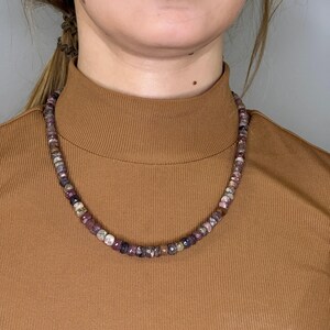 Op de afbeelding: Een ketting van veelkleurige edelstenen, met roze, paarse en witte tinten. De ketting is gemaakt van kleine, ronde kralen. Het model draagt een bruine coltrui. De ketting zit om de nek.