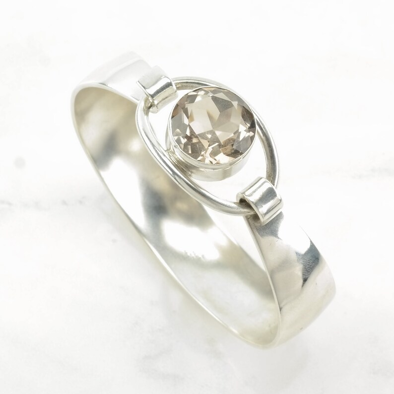 Peut inclure: Un bracelet jonc en argent avec une pierre ronde en quartz clair sertie dans un cadre en argent. Le bracelet est ouvert et a une finition lisse et polie.