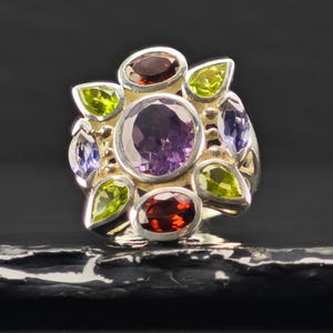 May include: A silver ring featuring a large central oval amethyst surrounded by a cluster of smaller faceted gemstones. The arrangement includes two deep red garnets, four bright green peridots, and two light purple tanzanite stones.