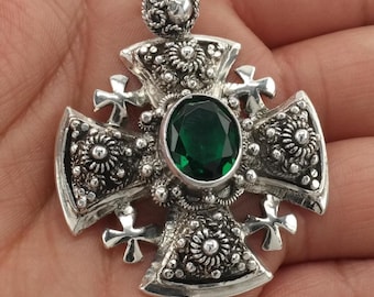 May include: A silver pendant in the shape of a cross, featuring an oval, emerald-green gemstone at its center. The cross has ornate floral and cross details. The pendant is held in a hand.