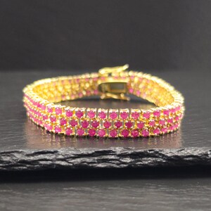 Pode incluir: Uma pulseira dourada com três fileiras de pedras preciosas rosa vibrantes. A pulseira possui um fecho seguro e é exibida em uma superfície escura e texturizada. As pedras preciosas são próximas, criando uma faixa contínua de cor.