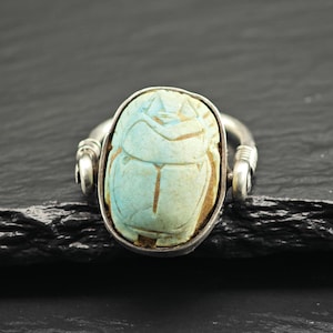 May include: A silver ring featuring a central, oval-shaped, light blue scarab beetle carving. The scarab has detailed, incised lines representing its wings and body. The metal band is secured to the sides of the setting with wire-wrapped loops.