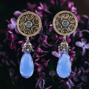 May include: A pair of ornate earrings featuring a gold-toned circular design with intricate scrollwork and a small blue gemstone center. Each earring has a silver-toned bell-shaped element and a large, faceted, teardrop-shaped blue gemstone pendant.
