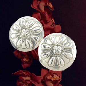 May include: Two silver-colored, round earrings with a floral design. Each earring features a central flower surrounded by petal-shaped elements. The earrings are set against a dark background with red orchid flowers.