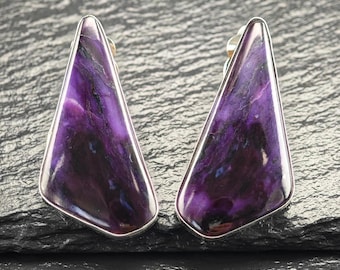 May include: A pair of triangular stud earrings. Each earring features a polished, vibrant purple stone with dark and light veining, set in a silver-toned metal frame. The earrings are displayed on a dark, textured surface.
