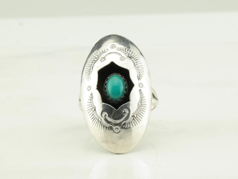 May include: A silver ring with an oval shaped setting. The setting has a turquoise stone in the center and a black background. The ring is decorated with a silver design around the edge.