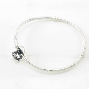 A silver bracelet with a charm on it is shown against a white marble background.