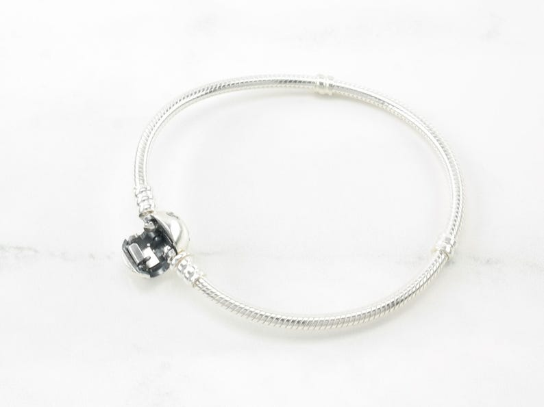 A silver bracelet with a charm on it is shown against a white marble background.