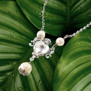 May include: A silver necklace with a heart-shaped pendant, adorned with a small red gemstone and three faux pearls. The necklace is set against a backdrop of large, green leaves with water droplets.