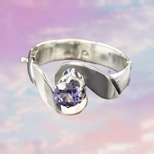 Op de afbeelding: Een zilveren ring met een grote, ronde, paarse edelsteen. De ring heeft een uniek, vloeiend ontwerp met een scharnierende sluiting. De edelsteen is gezet in een decoratieve zetting.