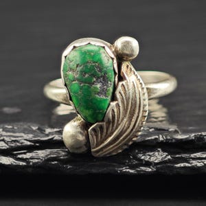 May include: A silver ring featuring a teardrop-shaped green turquoise stone with dark matrix patterns. The setting includes a polished silver leaf motif on one side, accented by two small silver spheres at the top and bottom of the stone.