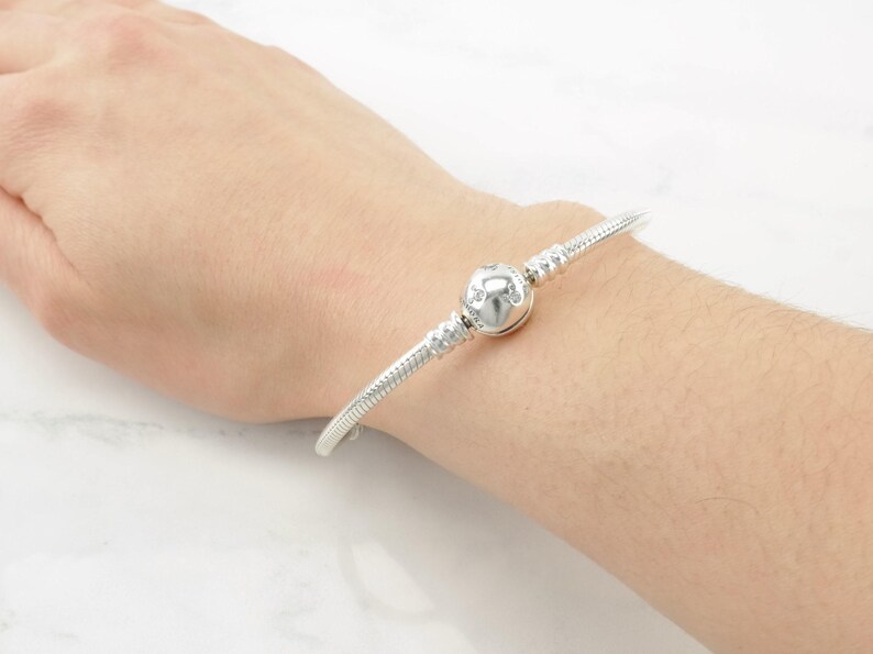 A silver bracelet with a round charm is worn on a person's wrist.