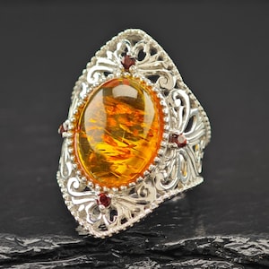 May include: Ornate silver ring featuring a large, oval amber gemstone with internal patterns. The ring has a filigree design with small red accent stones. The amber is a warm, golden color, set against a dark background.