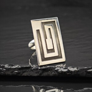 Puede incluir: Un anillo de plata con una cara rectangular que presenta un patrón geométrico en espiral empotrado y de tono oscuro. La superficie metálica es pulida y lisa, mostrando un diseño lineal limpio que se extiende por la parte superior plana.