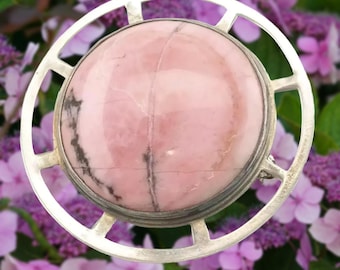 May include: A round, pink gemstone pendant with a silver-colored, openwork frame. The pink stone has darker veining and is set in a circular bezel. The frame has a geometric design, and the background is a blur of purple flowers.