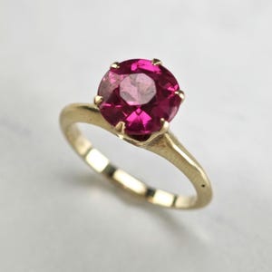 May include: A gold ring featuring a round, faceted, deep pink gemstone set in a six-prong mounting. The band is smooth and polished, showcasing a classic design that highlights the vibrant color and clarity of the central stone.