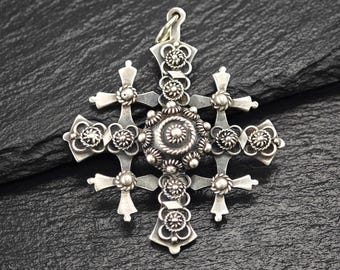 May include: Detailed silver pendant in the shape of a cross. The pendant features intricate floral and spiral designs, with smaller crosses at each arm. The pendant is displayed on a dark, textured surface.