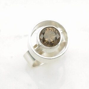 Peut inclure: Une bague en argent avec une pierre pr&eacute;cieuse ronde de couleur brune sertie dans un cadre circulaire. La bague a une bande simple.